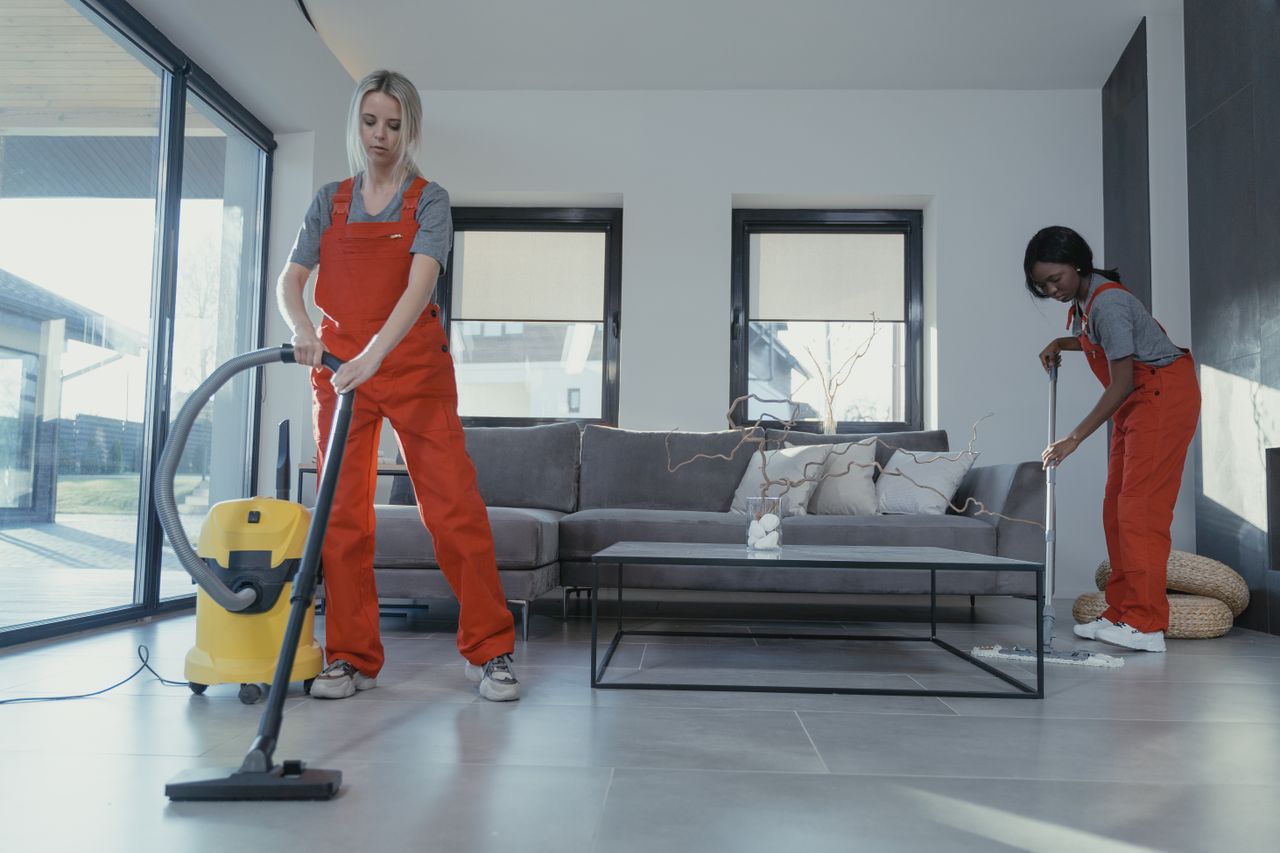 services-03 Two women in red overalls vacuuming and sweeping a modern living room.